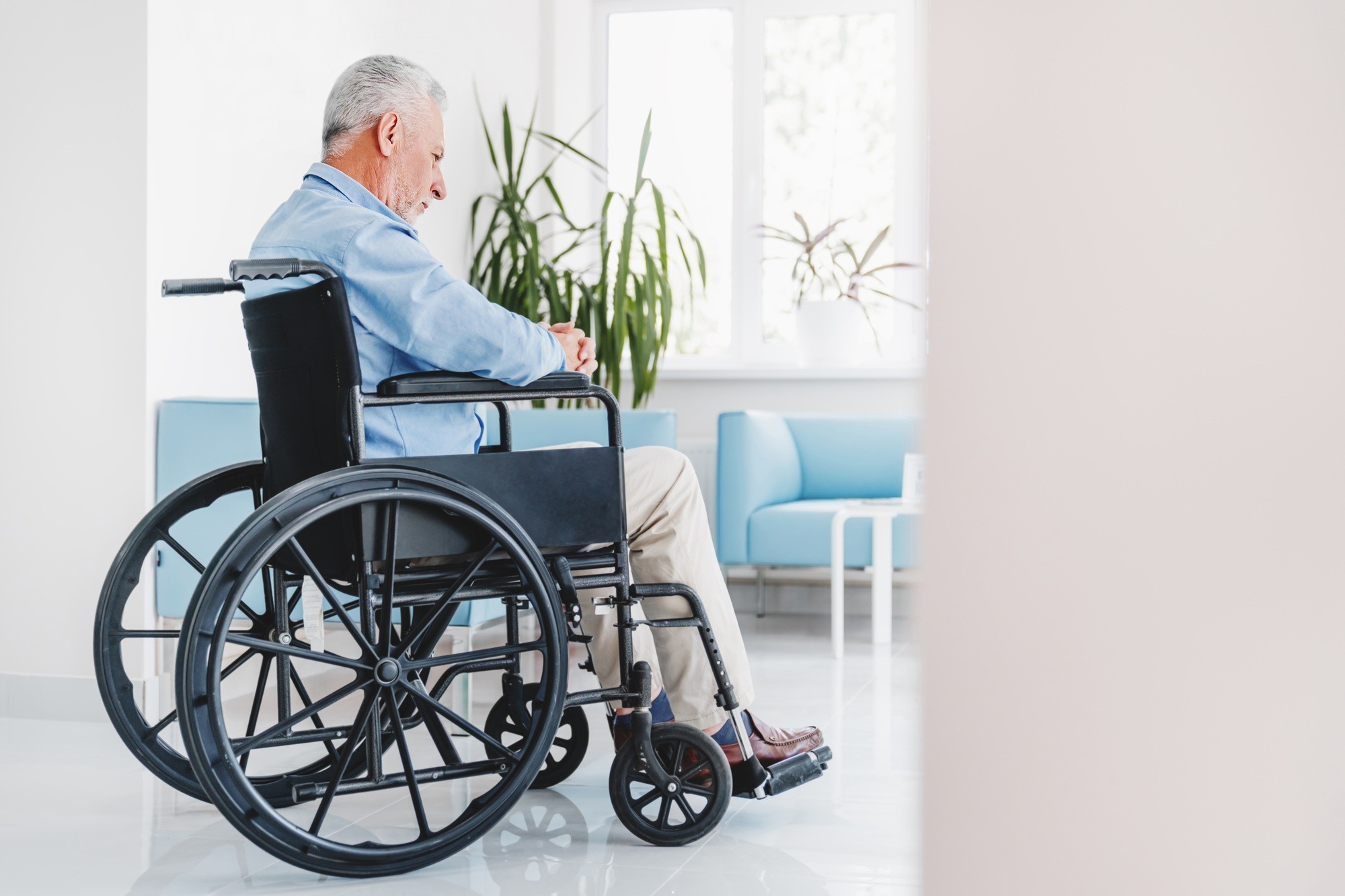 Sad elderly man sitting in wheelchair while looking out in window in retirement house